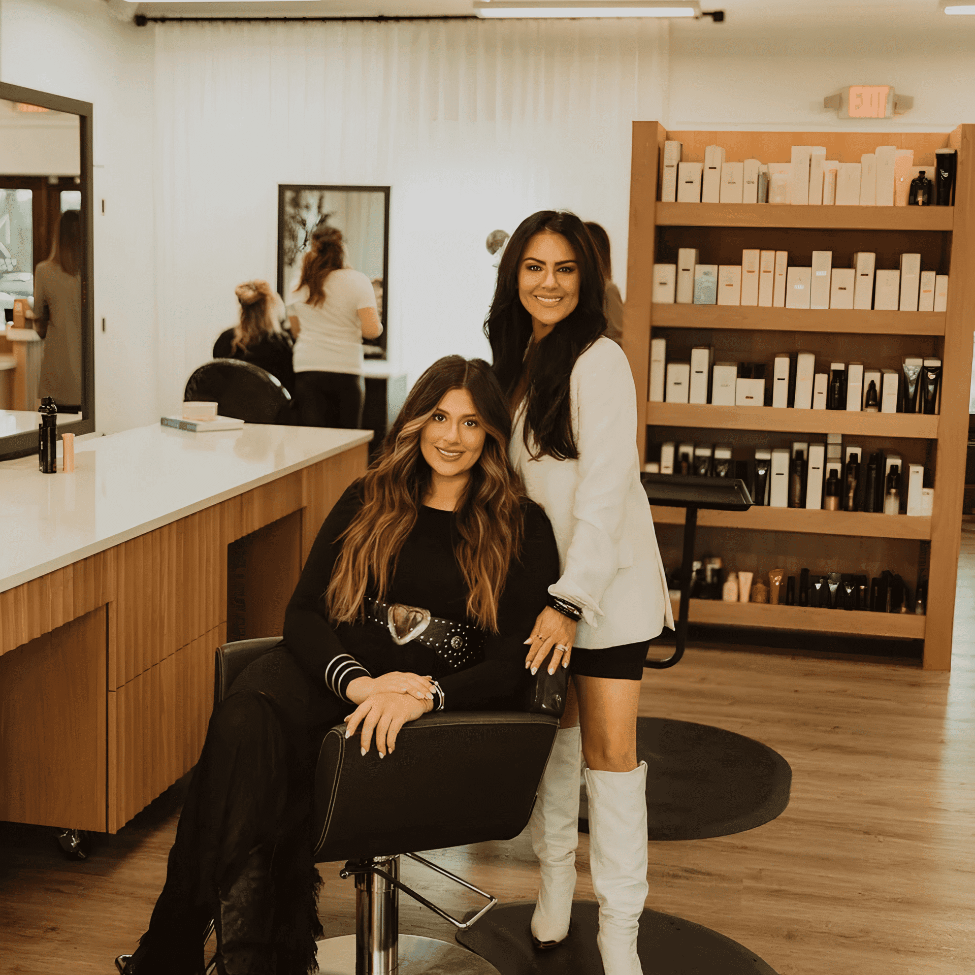Two women in a salon, one seated, one standing, shelves of beauty products in the background.