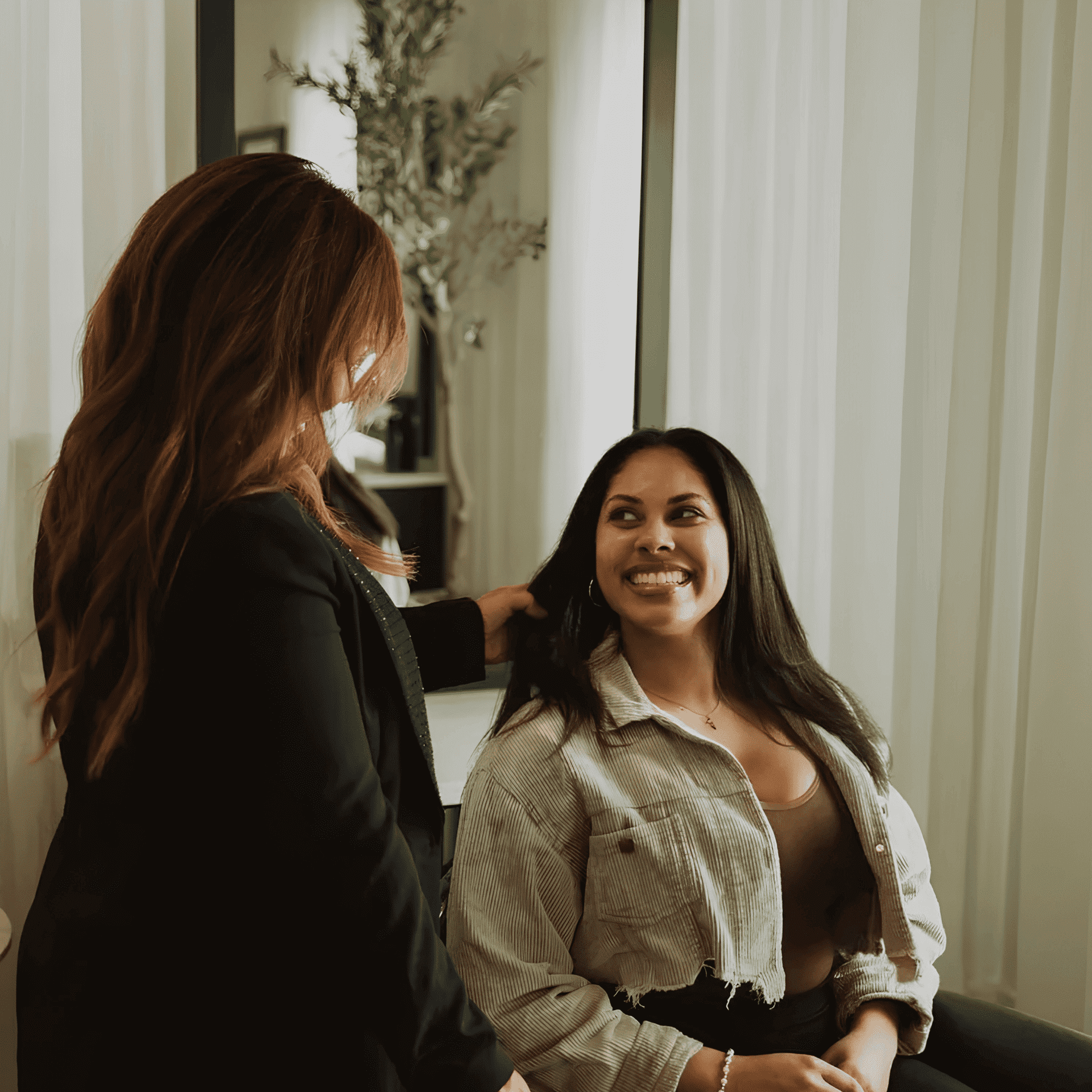 Two women smiling and talking in a modern room with plants and curtains.