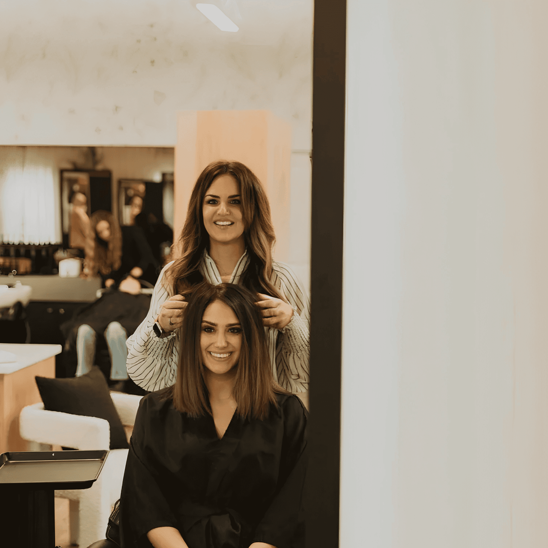 Hair stylist working on client's hair in salon, both smiling in mirror reflection.